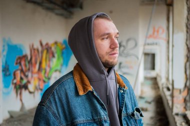 Portrait of young handsome guy in a denim jacket and a gray hood poses against the background of a destroyed and abandoned building with graffiti. Urban concept. Street lifestyle.