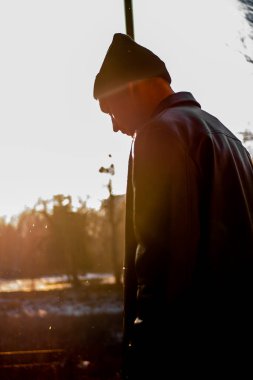 A handsome young guy in a black leather jacket and hat poses sideways with his head down against the background of the sun's rays. Urban concept. Vertical shot.