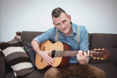 50 'li yaşlarda yakışıklı bir adam kanepede oturup gitar çalıyor.