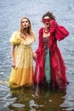 İki kadın, tarihi elbiseler içinde suyun içinde el ele tutuşuyorlar.