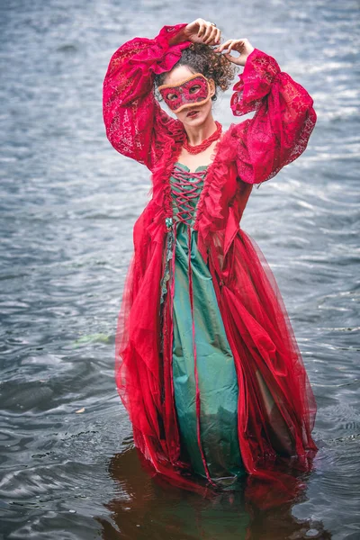 歴史的な赤いドレスを着たエレガントな女性と水中に立つ仮面舞踏会 — ストック写真