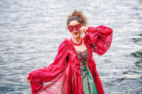 歴史的な赤いドレスを着たエレガントな女性と水のそばに立つ仮面舞踏会 — ストック写真