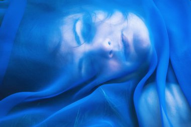 Çimenlerde uzanmış mavi şeffaf kumaşla genç bir kadının portresi.