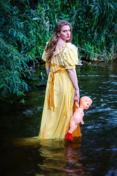 人形を持った水の中に立つ黄色の歴史的なドレスのブロンドの女性 — ストック写真