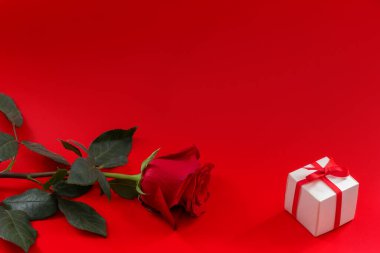 A gift box with a red ribbon and a rose flower on a red background. The concept of greetings for Valentine's Day, anniversary, Mother's Day and birthday, copy space, top view