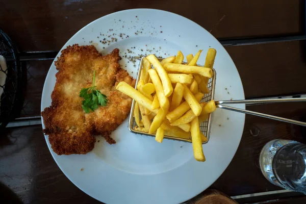 Polish breaded pork or chicken cutlet with coriander on top with fried potato chips or french fries. Polish meat cuisine on a white plate against wooden background
