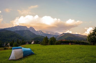 Gün batımında ve dramatik bulutlarda Tatra dağlarına karşı yeşil çayır arazisine kurulmuş kamp çadırı vahşi doğa kavramında macera.