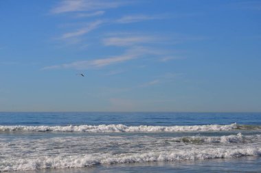 Pasifik Okyanusu 'nun deniz manzarası ve Gökteki Kuş. Mavi gökyüzü, Cardiff California plajı boyunca çakışan dalgaların üzerinde hafif bulutlarla. Ufuk düşük üçüncüde gökyüzünde fotokopi alanı var. Sahile zıt düşen dalgalar ön planda.