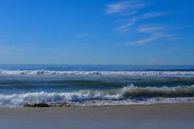 Mavi gökyüzü, Cardiff California plajı boyunca çakışan dalgaların üzerinde hafif bulutlarla. Ufuk, gökyüzünde fotokopi uzayının oluşmasına olanak sağlıyor. Sahile zıt düşen dalgalar ön planda.