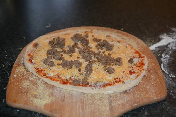 ピザを作る初期の段階の詳細を選択的に焦点を当てます 丸みを帯びたピザ生地とピザの皮は 生地が付着しないようにトウモロコシの食事で軽くほこりを消す ピザはソースチーズとソーセージで覆われています — ストック写真