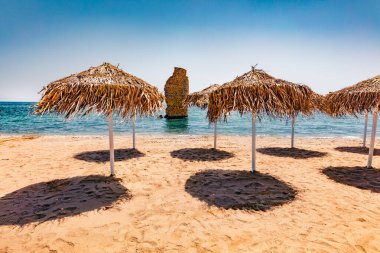 Arka planda güneş şemsiyeleri ve eski Roma kulesi kalıntıları olan boş Mylos Sahili. Peloponnese yarımadasının güneşli yaz manzarası, Voria Kinouria, Yunanistan, Avrupa. Parlak Myrtoanian deniz manzarası.