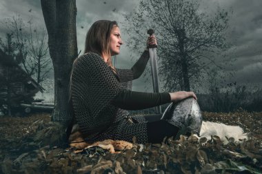 Dramatik gökyüzü altında ortaçağ savaşçısı suretinde bir kadın portresi. Ortaçağ zincirli güzel kadın savaştan sonra oturur.