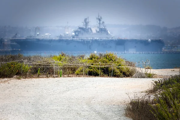 A gravel path on the Silver Strand, south of Coronado, California, leads to San Diego Bay with a US Navy Wasp-class assault ship (LHD) in the background.