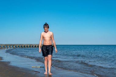 Şortlu genç sahilde yürüyor, yanında iskele var.