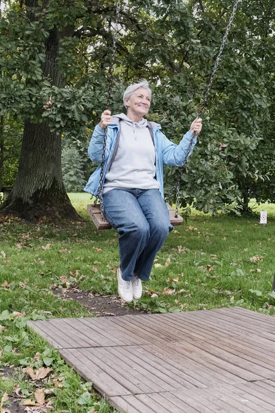 Happy senior caucasian woman with short gray hair swinging on chain swing in park — Stock Photo, Image