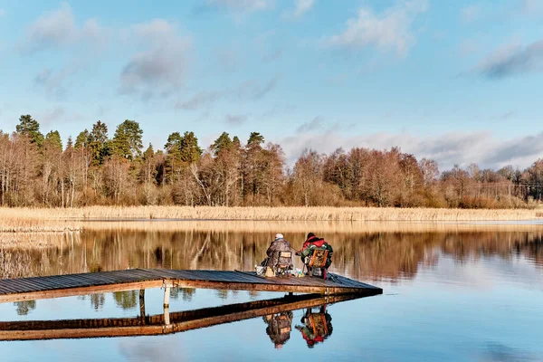 Dwóch rybaków z wędkami siedzących na drewnianym moście wśród malowniczego jeziora, odbijających się na lustrzanej powierzchni wody. — Zdjęcie stockowe