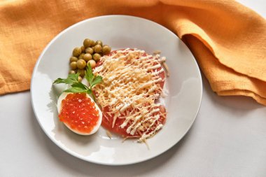 Haşlanmış tavuk yumurtasının yarısında tuzlu kırmızı balık havyarı, rendelenmiş sert peynirin altında taze kırmızı domates dilimleri ve beyaz masada mayonez şeritleri. Kahvaltı konsepti. Üst Manzara. Seçici odak.