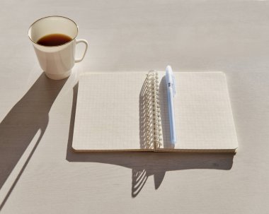 Open blank notebook and small white cup with coffee on white table in bright sunlight