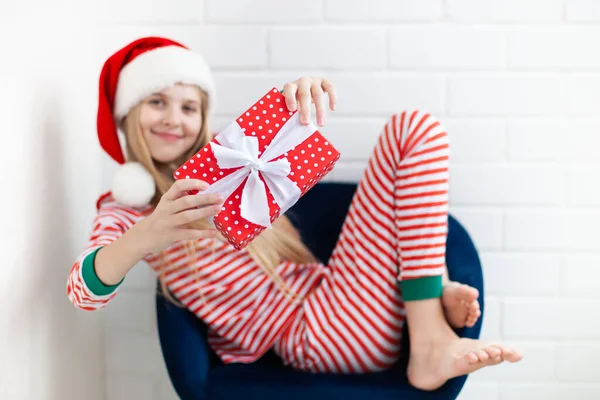 縞模様のパジャマとサンタクロースの帽子を身に着けている肯定的な女の子は白い弓でクリスマスのギフトボックスを保持する クリスマスとビューイヤーのコンセプト 選択的焦点 — ストック写真