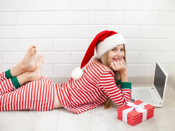 白いレンガの背景にラップトップを使用してストライプのパジャマとサンタクロースの帽子を身に着けている肯定的な女の子 クリスマスと新年のオンラインショッピング オンラインコースの販売の概念 選択的焦点 — ストック写真