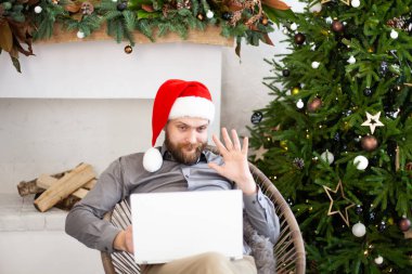 Kırmızı Noel Baba şapkası takan sakallı adam bilgisayarını kullanarak internet üzerinden arama yapıyor. Ev ofisinin rahat oturduğu web kamerasına merhaba diyor. Noel online tebrik konsepti. Seçici odak.