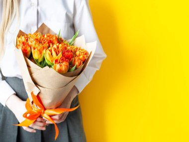 Close up of girl holding bouquet of bright orange tulips on a yellow background. Copy space. Spring holidays, mothers day, womens day, teachers day concept. Selective focus.