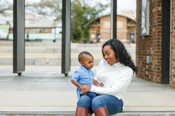 Красивая афро-американская мама сидит на ступеньках и обнимает своего сына-малыша, и они оба улыбаются. — стоковое фото