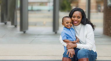 Güzel bir Afro-Amerikalı anne dışarıda merdivenlerde oturuyor ve yürümeye yeni başlamış oğluna sarılıyor ve ikisi de gülümsüyor ve mutlu.