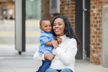 Güzel bir Afro-Amerikalı anne dışarıda merdivenlerde oturuyor ve yürümeye yeni başlamış oğluna sarılıyor ve ikisi de gülümsüyor ve mutlu.