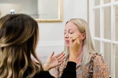 Kozmetik uzmanı, makyaj koltuğunda oturan sarışın bir kadına makyaj yapıyor.