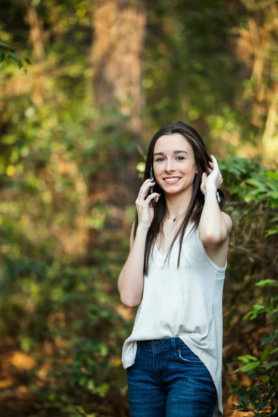 En vacker glad tonåring brunett flicka utanför på sin mobiltelefon i ett skogsområde på våren — Stockfoto