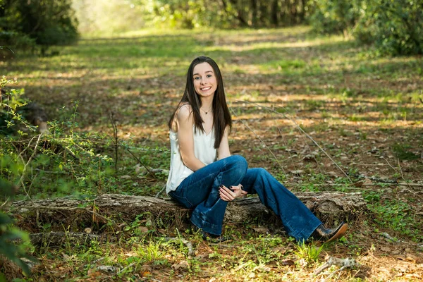 A beautiful happy teenage brunette girl sitting outdoors on a log hapy and smiling with copy space — Fotografia de Stock