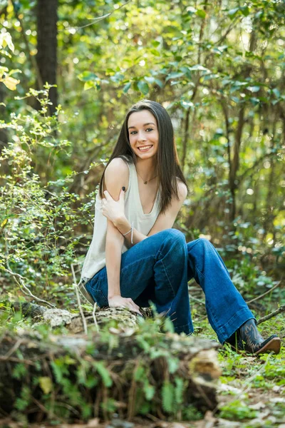 Une belle adolescente brune heureuse assise à l'extérieur sur une bûche hapy et souriante avec espace de copie — Photo