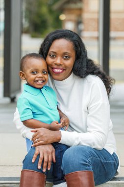 Güzel bir Afro-Amerikalı anne dışarıda merdivenlerde oturuyor ve yürümeye yeni başlamış oğluna sarılıyor ve ikisi de gülümsüyor ve mutlu.