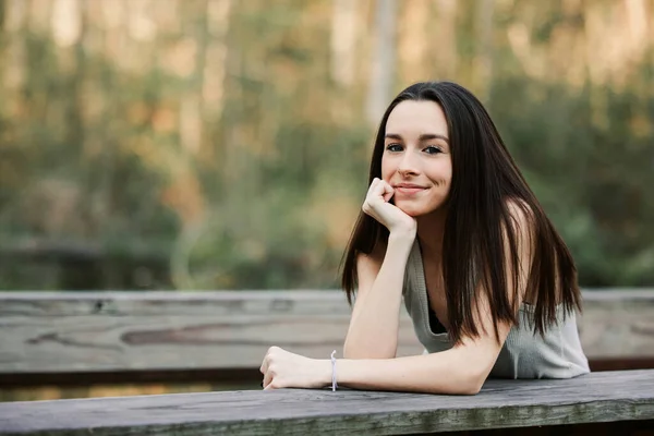A tall and thin brunette teenager standing and leaning on a park bridge with her head resting on her hands with a smile and joyful expression — Zdjęcie stockowe