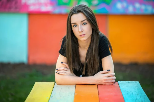 A beautiful serious teen girl leaning forward on a vibrant multi-colored striped picnic table with a bright background — Zdjęcie stockowe
