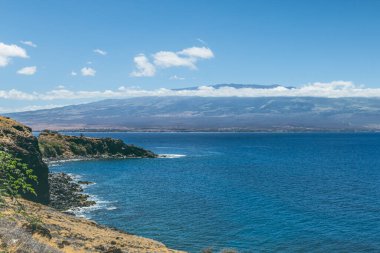 A high angle sweeping view of Maui Hawaii at mid day with a mountain, clouds and the Pacific ocean in the distance.