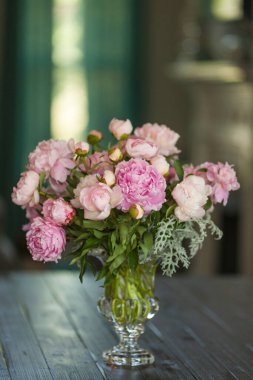 Bir buket yumuşak pembe şakayık, cam bir vazoda kahverengi ahşap bir kırsal masada oturuyor..