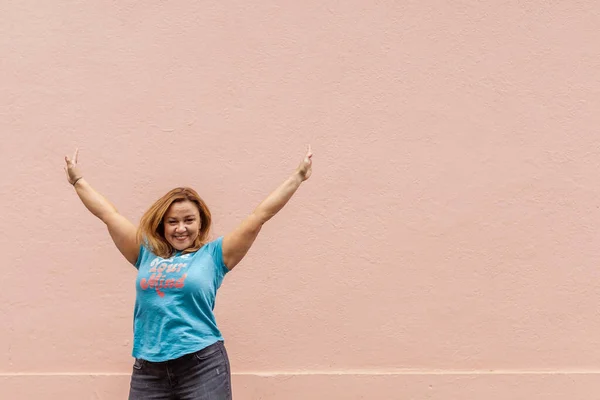 waist up of a woman in jeans and jumping with her arms raised, against a peach exterior wall background outside.