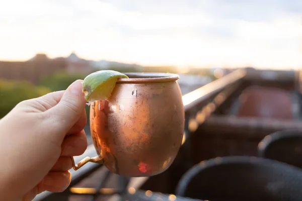 A moscow mule with a lime held up at a rooftop restaurant bar at sunset.