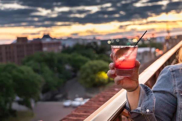 A hand holding a red pink rasberry alcoholic beverage at a Savannah, Georgia at sunset at a rooftop bar.
