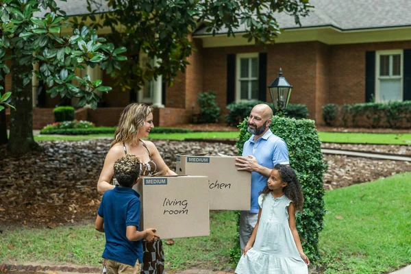 A blended family mom and dad and two young children holding packing boxes and moving into their new home.
