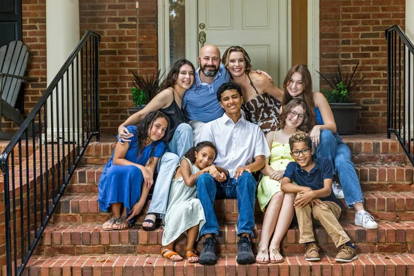 A large and blended and mixed happy family sitting on the front steps of their brick home in the suburbs.