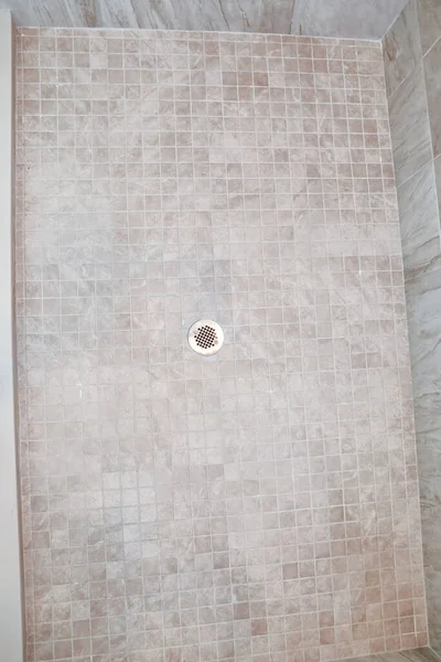 A neutral light beige square tile shower floor with a silver metal drain.