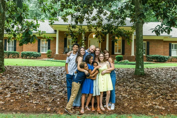 A large and blended and mixed happy family standing in front of their home in a group hug or embrace.