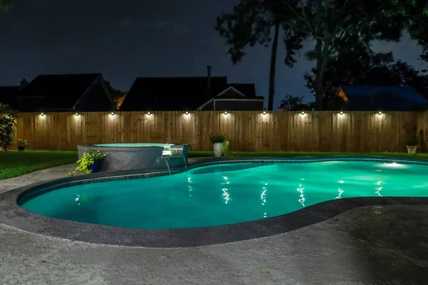 A backyard swimming pool and jacuzzi hot tob at night with solar lights around the fence for privacy and illumination.