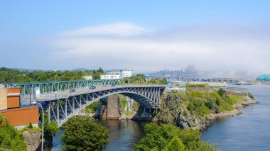 Saint John, NB, Kanada 'daki Reversing Falls köprüsü, Wolastoq Park' tan görüldü. Sis uzaktaki şehir merkezinin üzerinde süzülüyor. Güneşli bir gün.