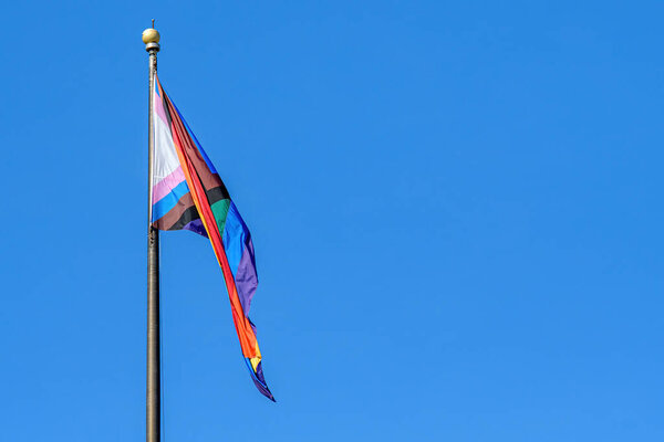 A LGBTQ+ rainbow progress pride flag hangs limply on a flag pole. Blue sky behind.