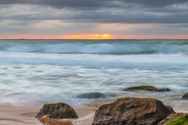 Kumsalda kayalarla birlikte gün doğumu denizi ve Orta Sahildeki Killcare Sahili 'nden gelen alçak bulutlar, NSW, Avustralya.