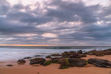 Kumsalda kayalarla birlikte gün doğumu denizi ve Orta Sahildeki Killcare Sahili 'nden gelen alçak bulutlar, NSW, Avustralya.